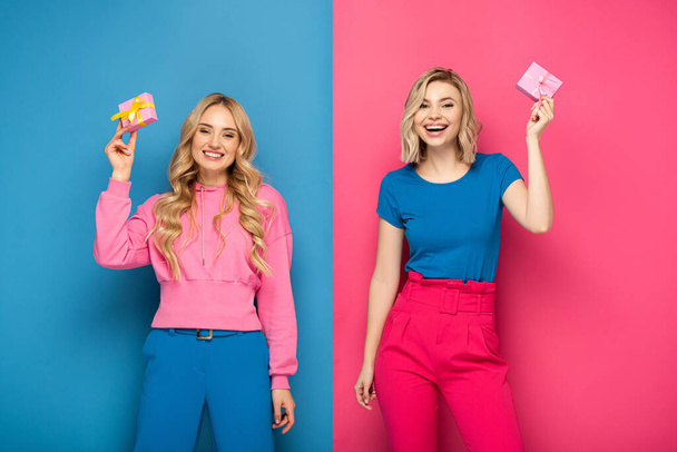 glimlachende blonde meisjes met geschenken en kijken naar de camera op blauwe en roze achtergrond - Foto, afbeelding