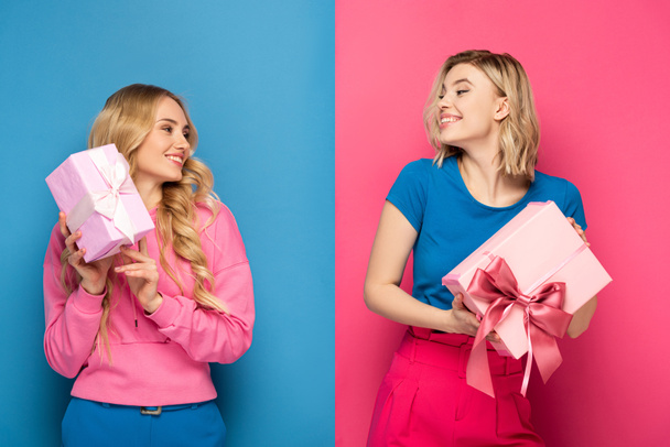 Smiling blonde girls looking at each other while holding gift boxes on blue and pink background - Φωτογραφία, εικόνα