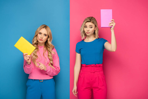 blond meisjes kijken naar camera terwijl het houden van boeken over blauw en roze achtergrond - Foto, afbeelding