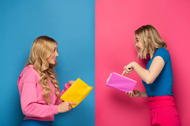 Zijaanzicht van glimlachende blonde zussen kijken naar elkaar terwijl het houden van boeken over blauwe en roze achtergrond - Foto, afbeelding