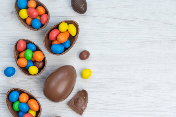Chocolate Easter eggs and chocolate eggs filled with sweet and colorful candies, on white wood table. Copy space , top view. - Fotó, kép