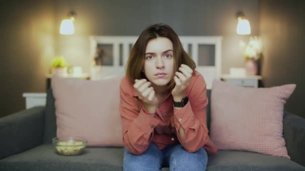 Young enthusiastic woman sitting on the sofa, eating popcorn and watching TV - Filmati, video