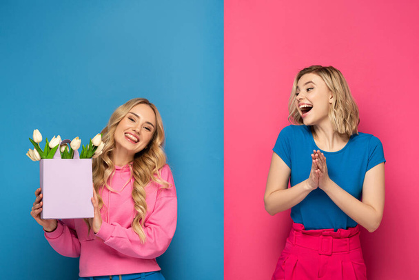 Cheerful blonde sister with bouquet in shopping bag on pink and blue background - Foto, Imagem