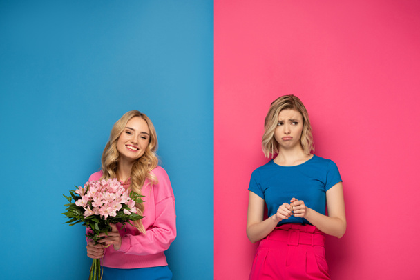 Beautiful blonde girl smiling while holding bouquet near offended sister on pink and blue background - Фото, зображення