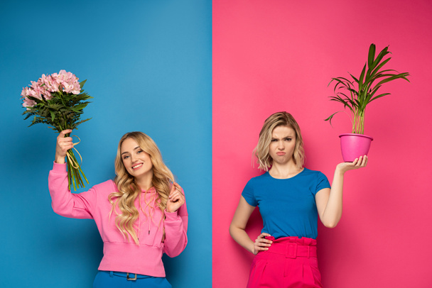 Smiling girl holding bouquet near offended sister with plant on pink and blue background - Fotó, kép