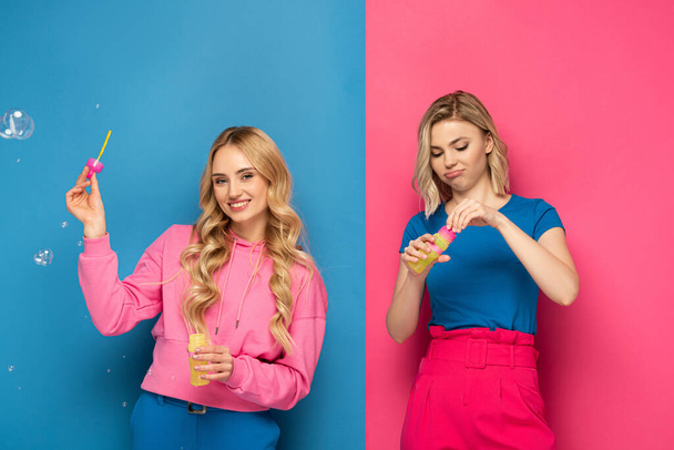 Lächelndes blondes Mädchen mit Seifenblasen in der Nähe der Schwester auf rosa und blauem Hintergrund - Foto, Bild