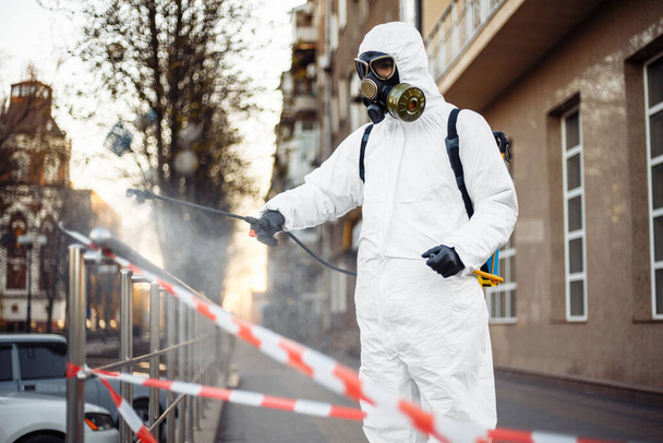 Ein Mann in Schutzausrüstung desinfiziert sich in der Stadt mit einem Sprayer. Oberflächenbehandlung wegen Coronavirus covid-19 Krankheit. Ein Mann im weißen Anzug desinfiziert Straße und Schienen. Virus-Pandemie - Foto, Bild