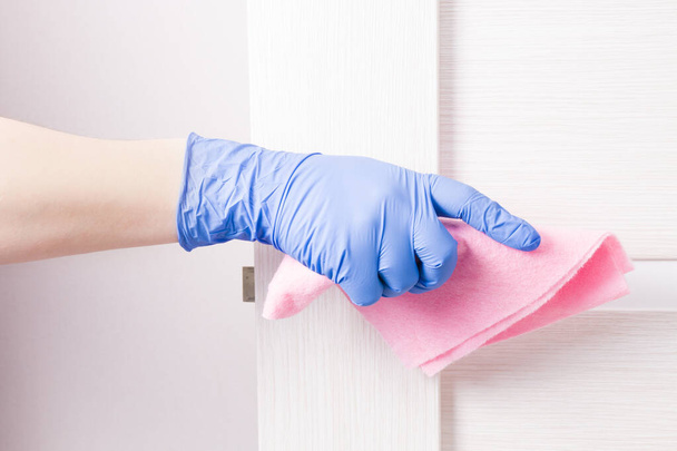 a hand in a blue disposable rubber glove wipes a doorknob with a pink rag, cleaning and disinfecting surfaces, the safety of your home, copy space, metal handle, light wooden door - Fotografie, Obrázek