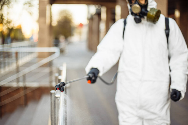 Man sprays disinfector onto the railing wearing coronavirus protective suit and equipment. Cleaning and sterilizing the not crowded city streets. Covid-19 nCov2019 spread prevention - Photo, Image