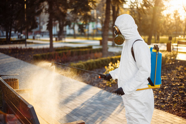 Özel korumalı dezenfeksiyon kıyafeti giyen bir adam şehirde yayılan koronavirüsün ortasında boş bir parktaki bankın üzerine sterilizatör sıkar. Güneşli bir arka plan. Dünya çapında Covid-19 'u durdurun. - Fotoğraf, Görsel