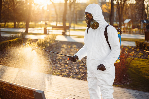 Egy férfi, aki speciális védőruhát visel, sterilizálót permetez egy padra az üres parkban, a városban elterjedt koronavírus közepére. Napfényes háttér. Stop Covid-19 világszerte - Fotó, kép