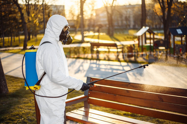 Человек в специальном защитном костюме дезинфекции распыляет стерилизатор на скамейке в пустом парке на фоне распространения коронавируса в городе. Солнечный фон. Stop Covid-19 повсюду
 - Фото, изображение
