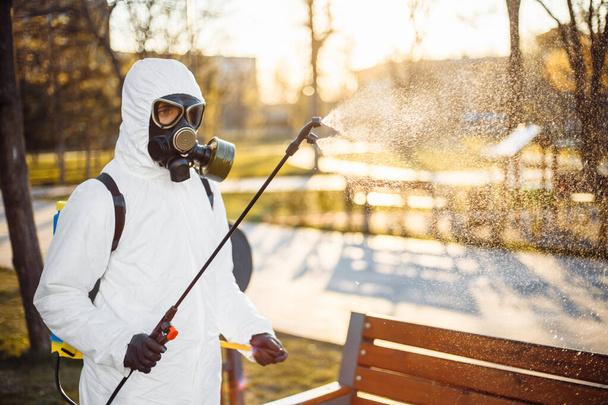 専用の防疫服を着た男が、市内に広がるコロナウイルスの中に、空の公園のベンチに滅菌器をスプレーします。晴れた背景。世界中でCovid-19を停止 - 写真・画像