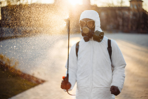 Koruyucu dezenfeksiyon kıyafeti giyen bir adam koronavirüs covid-19 'un yayılmasını önlemek için park meydanına sterilizatör sıkar. Profesyonel temizlik işçisi şehrin sokaklarını virüsten temizliyor. Nokta no. Cov2019 - Fotoğraf, Görsel
