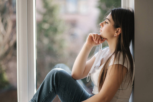 Young woman sits on windowsill and hold protective mask in hand. Female and look out the window. Alone at home in quarantine. Coronavirus theme. Stay at home - Foto, Imagem