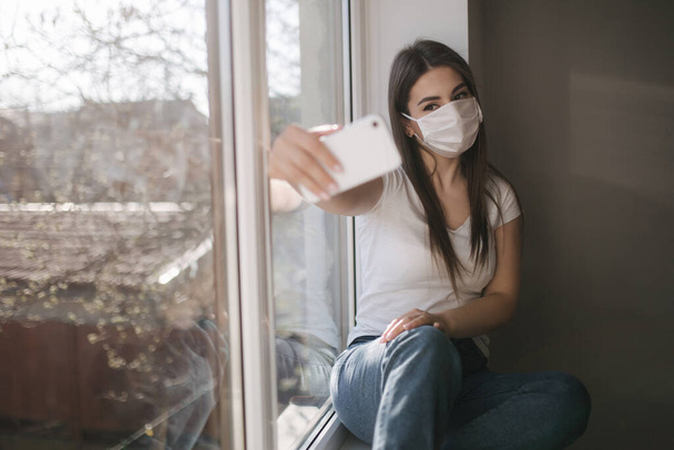 Aantrekkelijke vrouw met medisch masker maken selfie thuis. Vrouw in wit tshirt en wit medisch masker gebruik telefoon - Foto, afbeelding