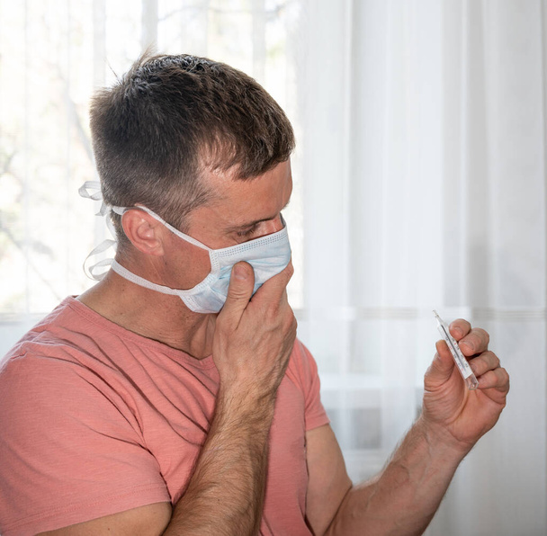 Gesundheitswesen und medizinisches Konzept. Mann mit Mundschutz hält Thermometer zu Hause - Foto, Bild