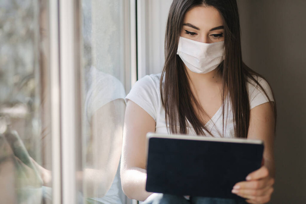 Attraktive junge Frau in weißer medizinischer Maske sitzt auf dem Fenstersims und bedient sich eines Tablets. Weibchen ruhen sich zu Hause in Quarantäne aus - Foto, Bild