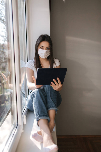 Attraente giovane donna in maschera medica bianca si siede sul davanzale della finestra e utilizzare tablet. Le donne hanno un riposo a casa in quarantena
 - Foto, immagini