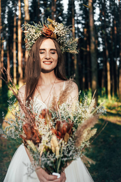 beautiful fabulous happy brunette bride with stylish bouquet and wreath. stylish bride looks at boho bouquet on background of forest, luxury gorgeous boho wedding at forest outdoors. - Foto, Bild