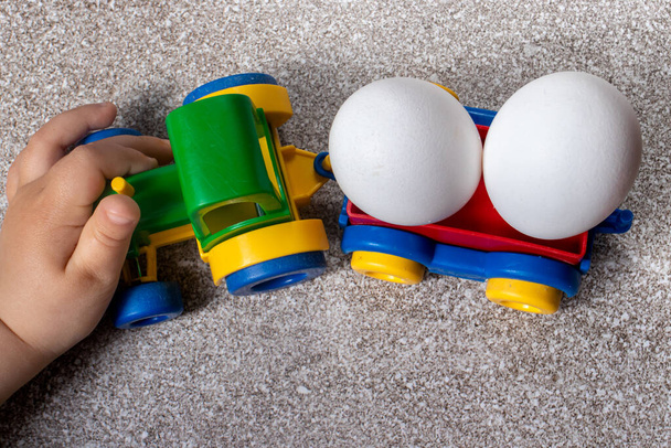 Ein kleines Kind spielt mit einem Spielzeugtraktor, der zwei Hühnereier transportiert. Plastik und natürliche Lebensmittel. - Foto, Bild