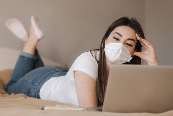 Jonge vrouw met masker gebruiken laptop op het bed. Vrouwelijke bekijk online video tijdens quarantaine in de slaapkamer. Studeren thuis - Foto, afbeelding
