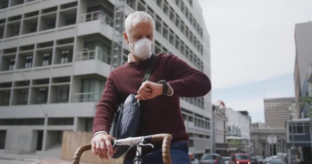Middle aged Caucasian man out and about in the city streets during the day, wearing a face mask against coronavirus, covid 19, sitting on his bicycle and using his smartwatch, in slow motion. - Footage, Video