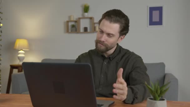 A white man with a beard sits in a room at a table, communicates on the Internet, looks at the interlocutor through the screen and yawns, in a hurry - Materiał filmowy, wideo
