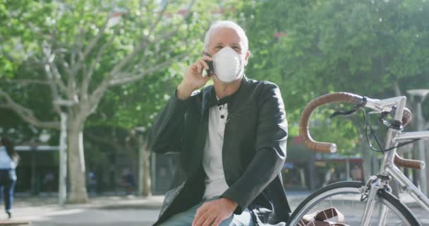 Middle aged Caucasian man out and about in the city streets during the day, wearing a face mask against coronavirus, covid 19, sitting on stairs and talking on his smartphone, in slow motion. - Felvétel, videó
