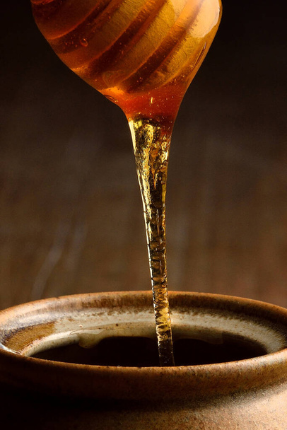 Μέλι ρίχνει από ξύλινο μέλι dipper σε κεραμική κούπα. Βάζο επίγειων σκευών για μέλι σε ξύλινο τραπέζι. - Φωτογραφία, εικόνα