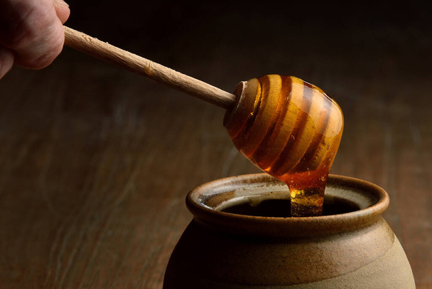 Honig, der aus einem hölzernen Honiglöffel in einen Keramikbecher fließt. Tongefäß für Honig auf Holztisch Hand hält Löffel. - Foto, Bild