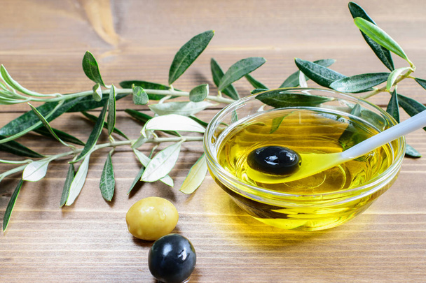 Черная оливка в ложке и прозрачная стеклянная чаша с оливковым маслом. Немного оливок и листьев на деревянном фоне. Здоровое питание
.  - Фото, изображение