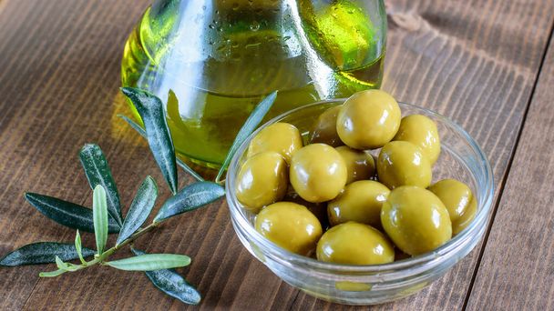 Консервированные зеленые оливки в прозрачной стеклянной миске и бутылке оливкового масла на деревянном столе кухни. Здоровое питание
.  - Фото, изображение