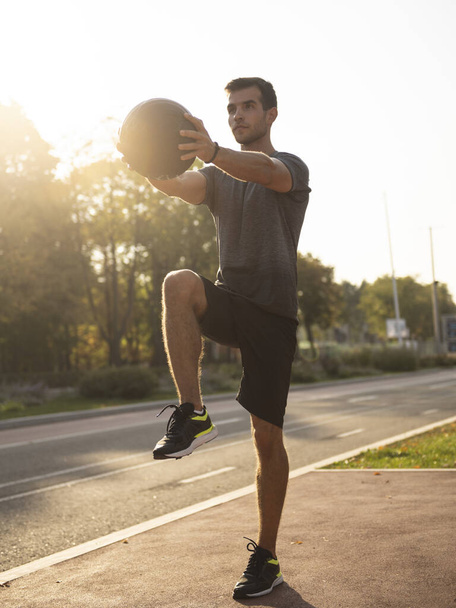 Fit guy doing exercises using a ball outdoors. Young athletic man training in city park - Photo, Image