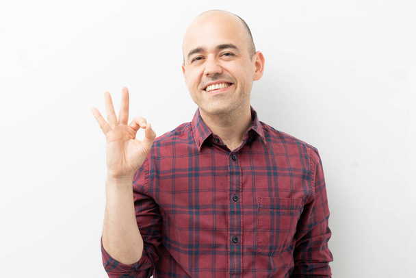 Kale man in de 30 maakt een goed teken met zijn hand en glimlacht in een studio - Foto, afbeelding
