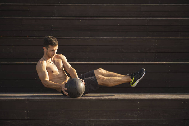 Fit guy doing abdominal exercise on a bench using a ball outdoor. Young athletic man training in city park - Фото, изображение