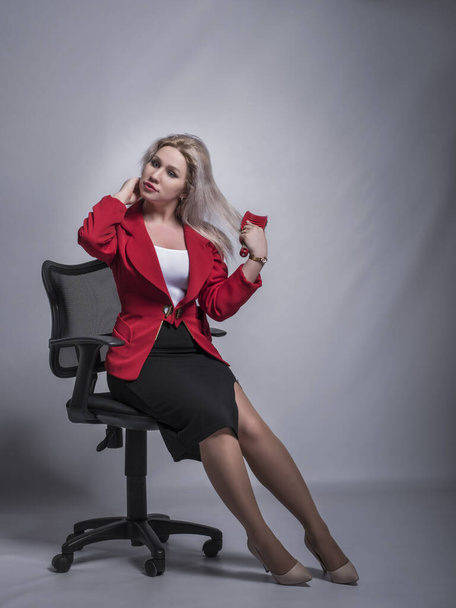Beautiful young girl, blonde, business woman in a red jacket and suit in an office chair on a gray and light background. - Photo, Image
