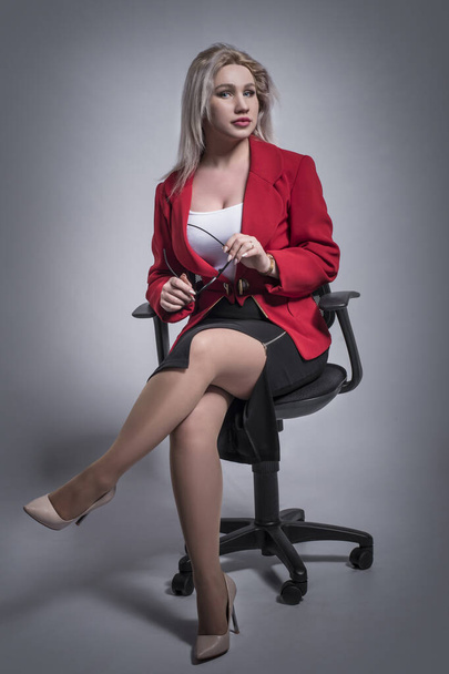 Beautiful young girl, blonde, business woman in a red jacket and suit, with glasses in an office chair on a gray and light background. - Fotografie, Obrázek