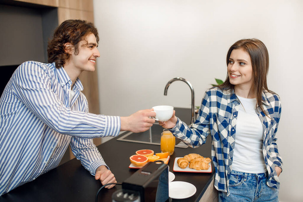 Portret van een gelukkig jong paar verliefd met behulp van koffiemachine, terwijl ze lekker ontbijten aan de tafel in een moderne licht keuken. - Foto, afbeelding