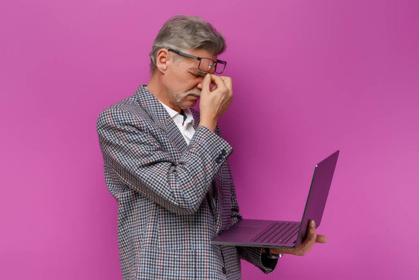 Tired old man holding laptop while staying on violet background. - Φωτογραφία, εικόνα