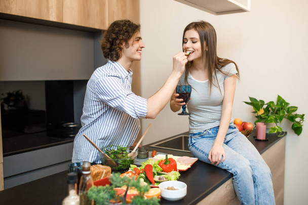 Young couple at modern kitchen, woman holding wine glass, male feeding woman. - Foto, imagen