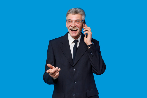 People lifestyle concept. Holding mobile phone elderly gray-haired mustache bearded man in classic suit posing isolated on blue background in studio. - Foto, Imagem