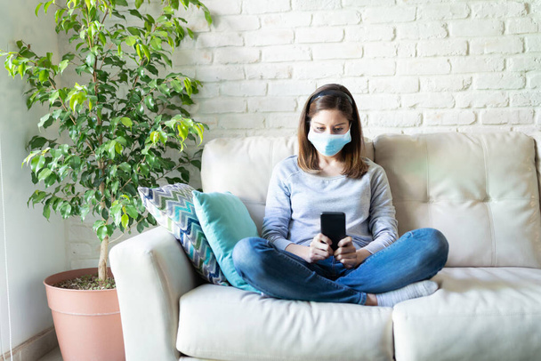 Aantrekkelijke brunette vrouw met behulp van een smartphone thuis tijdens het dragen van een chirurgisch masker en thuisblijven tijdens de gezelligheid19 pandemie - Foto, afbeelding