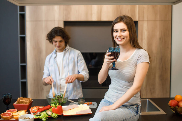 Young couple at kitchen, woman holding wine glass, male cooking salad with fresh vegetables. - Foto, Bild