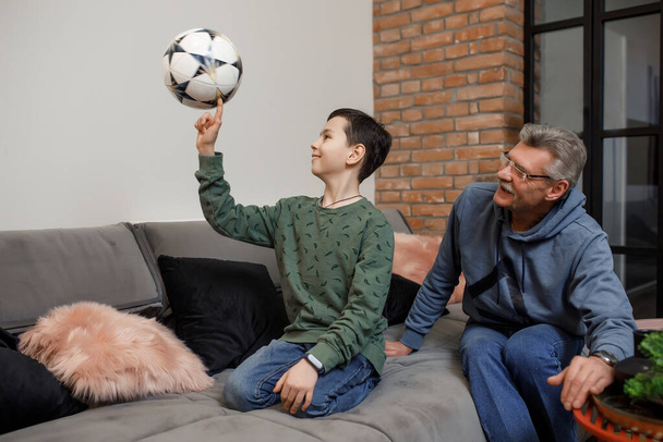 Smiling grandson playing with foootball ball with his happy gray grandfather while they sit on sofa. - Foto, immagini