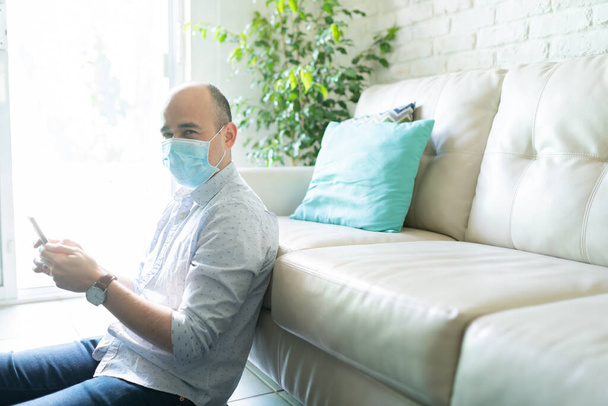 Retrato de um homem de 30 anos usando uma máscara cirúrgica em casa e usando seu smartphone e verificando as mídias sociais durante a pandemia de covid19
 - Foto, Imagem