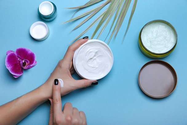 woman's hand takes some cream from a jar. Moisturizing cream in woman's hands on blue background with cosmetic jars flat lay. - Фото, изображение