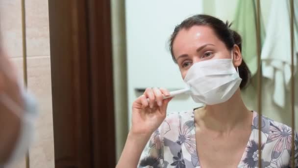 Attractive brunette woman in floral pajamas wear blue protective medical face mask, brush her teeth, look in mirror in bathroom close up. COVID-19 or corona virus year. Lady during epidemic situation. - Filmagem, Vídeo