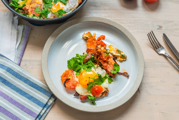 Frisches rustikales Frühstück - Spiegelei mit Speck, Zwiebeln und Kirschtomaten auf einem Teller auf hellem Holzgrund - Foto, Bild