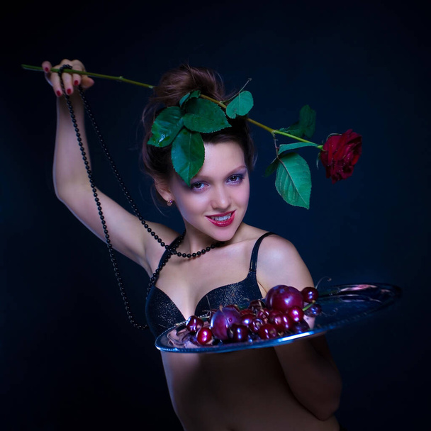 Conceptual portrait of a fashionable young, seductive, emotional, beautiful girl, woman in beautiful black underwear on a dark background, who is holding a tray with fruit. - Valokuva, kuva